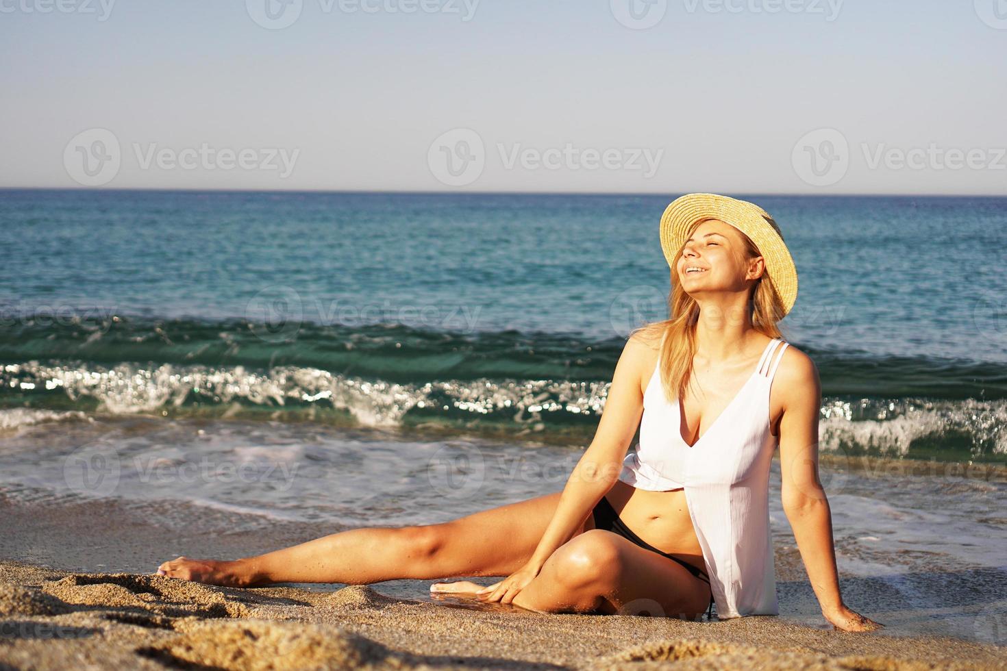Mädchen mit Strohhut und weißem Badeanzug sitzt auf einem Sand am Meer foto