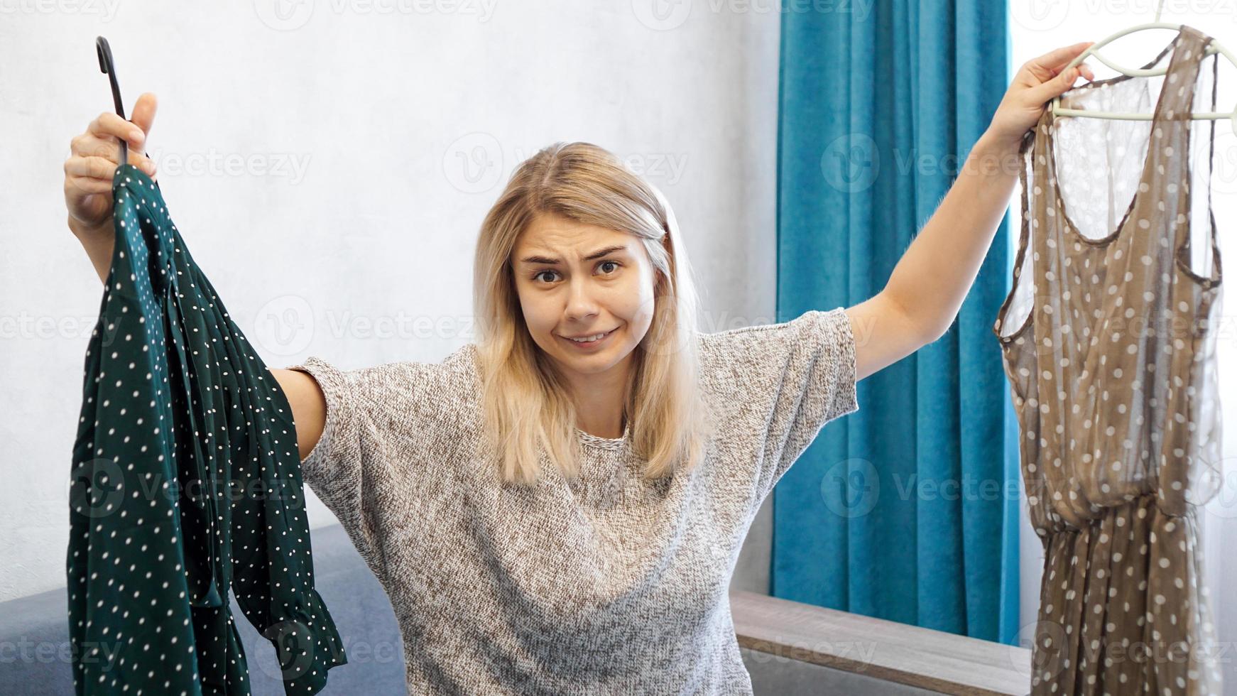 süße Frau, die ihr Outfit auswählt und zwei Kleider auf Kleiderbügeln hält foto