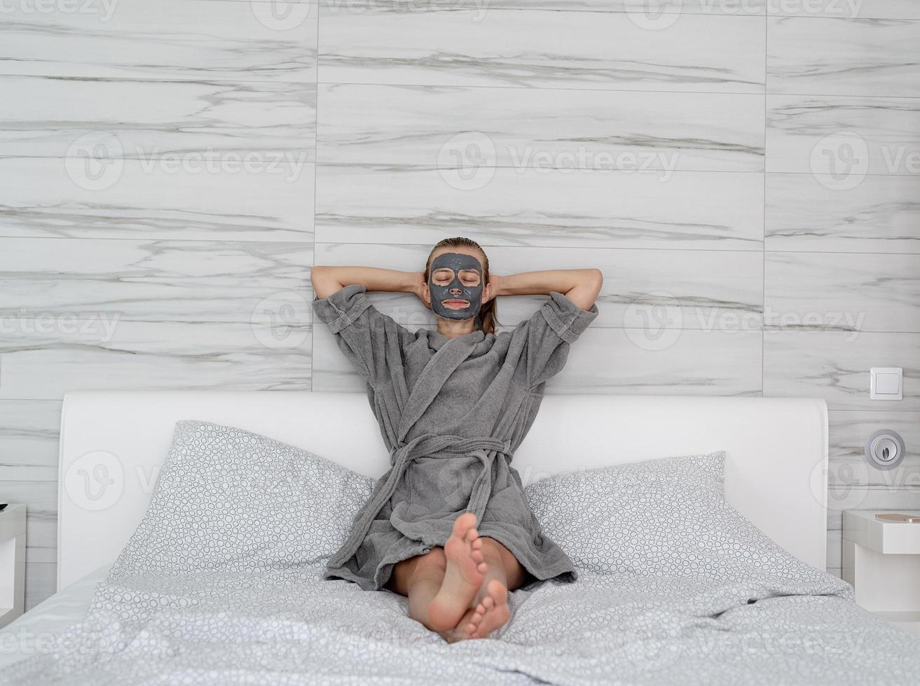 Frau mit Gesichtsmaske entspannt auf dem Bett sitzend foto