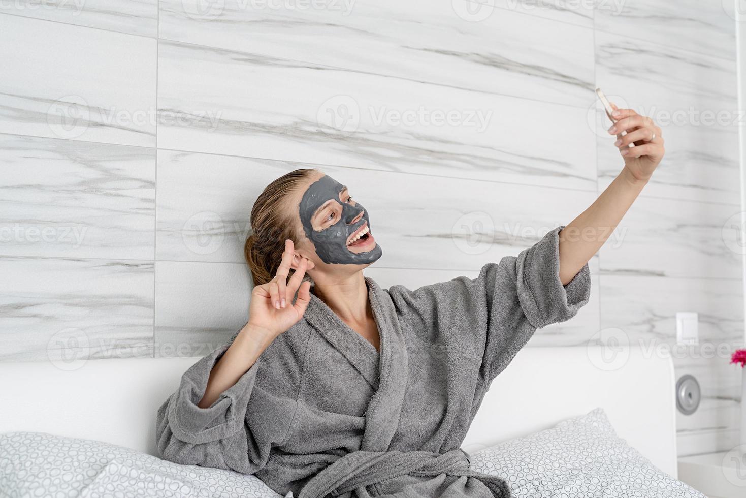 Frau mit Gesichtsmaske entspannt auf dem Bett sitzend mit einem mobilen Gerät foto