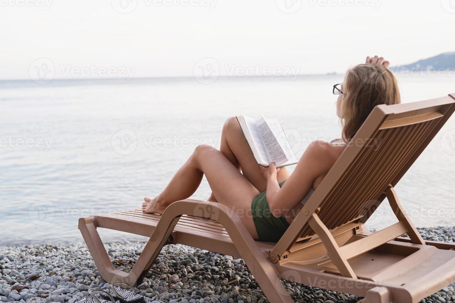 Die schöne junge Frau liegt auf der Sonnenliege und liest ein Buch foto