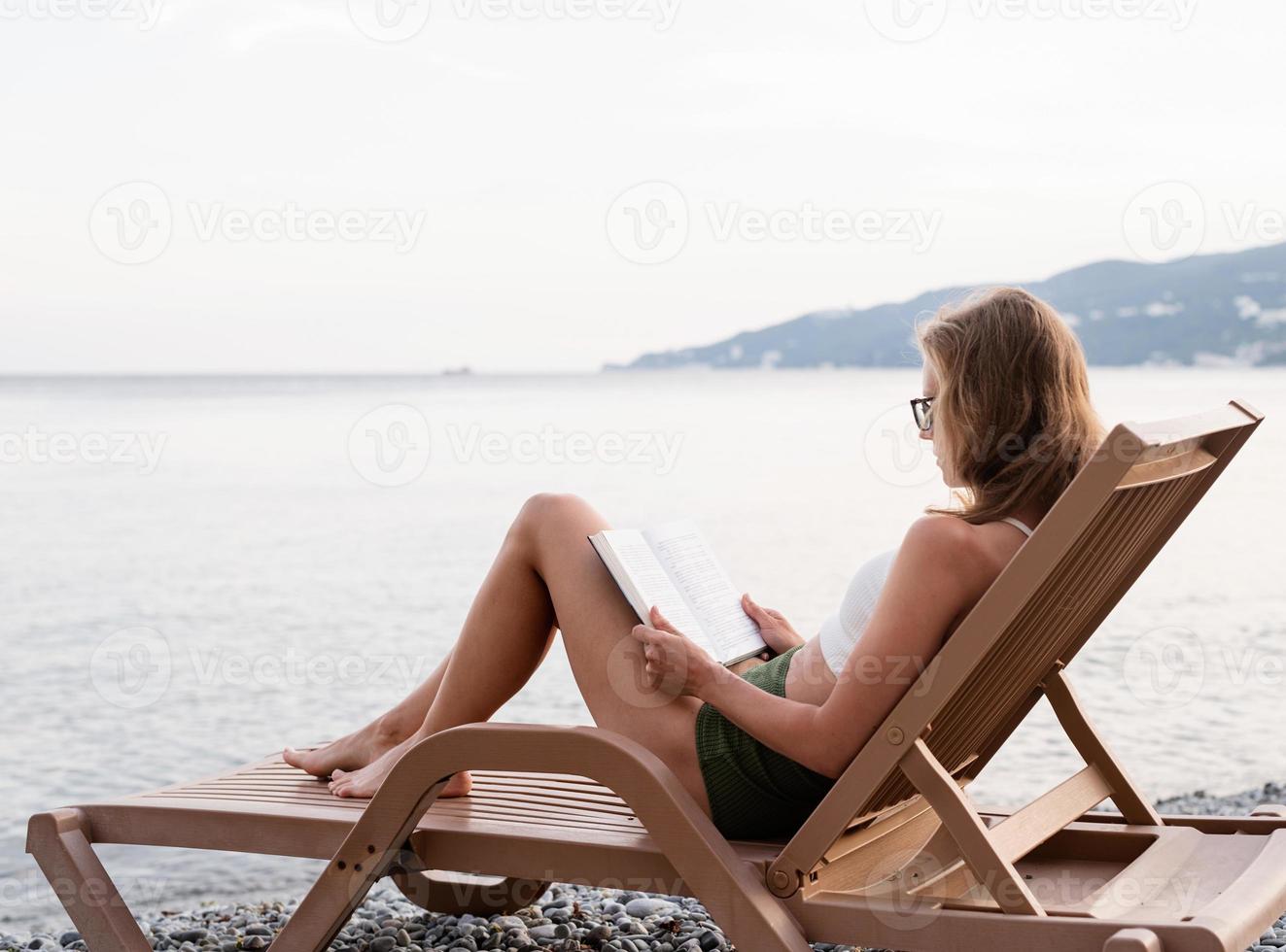 Die schöne junge Frau sitzt auf der Sonnenliege und liest ein Buch foto