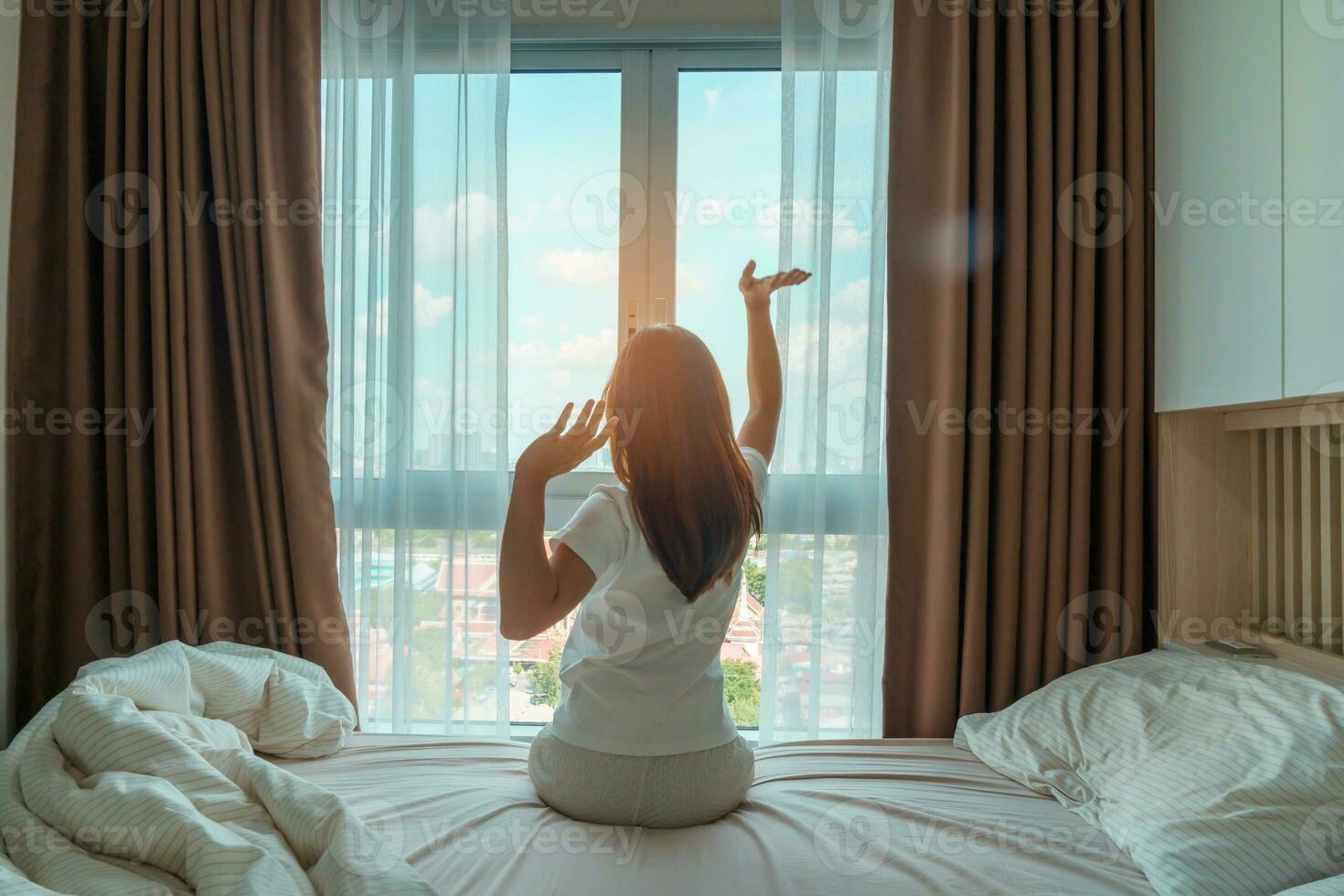 glücklich Frau Dehnen auf Bett nach aufwachen hoch, jung Erwachsene weiblich steigend Waffen und suchen zu Fenster im das früh Morgen. frisch entspannen und haben ein nett Tag Konzepte foto