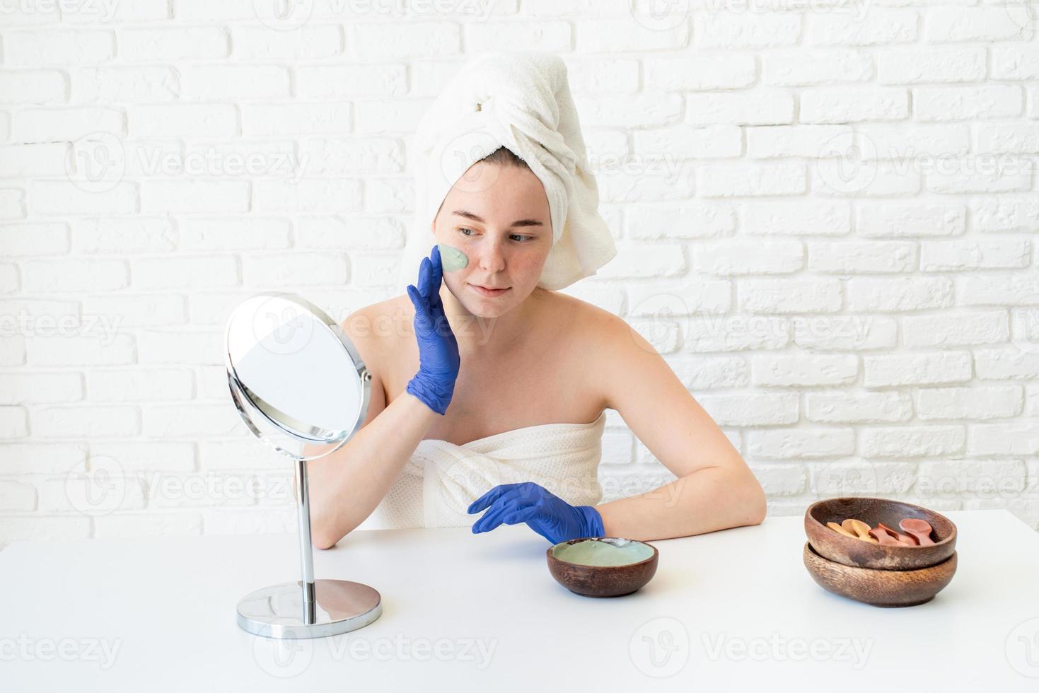 Frau mit Handschuhen, die eine Gesichtsmaske aus Ton anwendet und in den Spiegel schaut foto