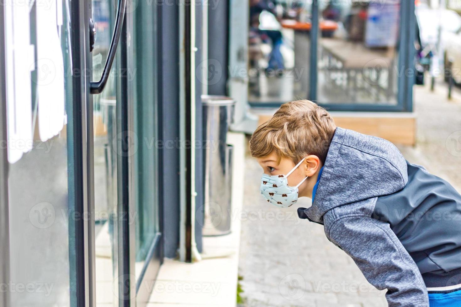 kleiner Junge mit Gesichtsmaske mit Blick auf das Schaufenster in der Stadt. foto