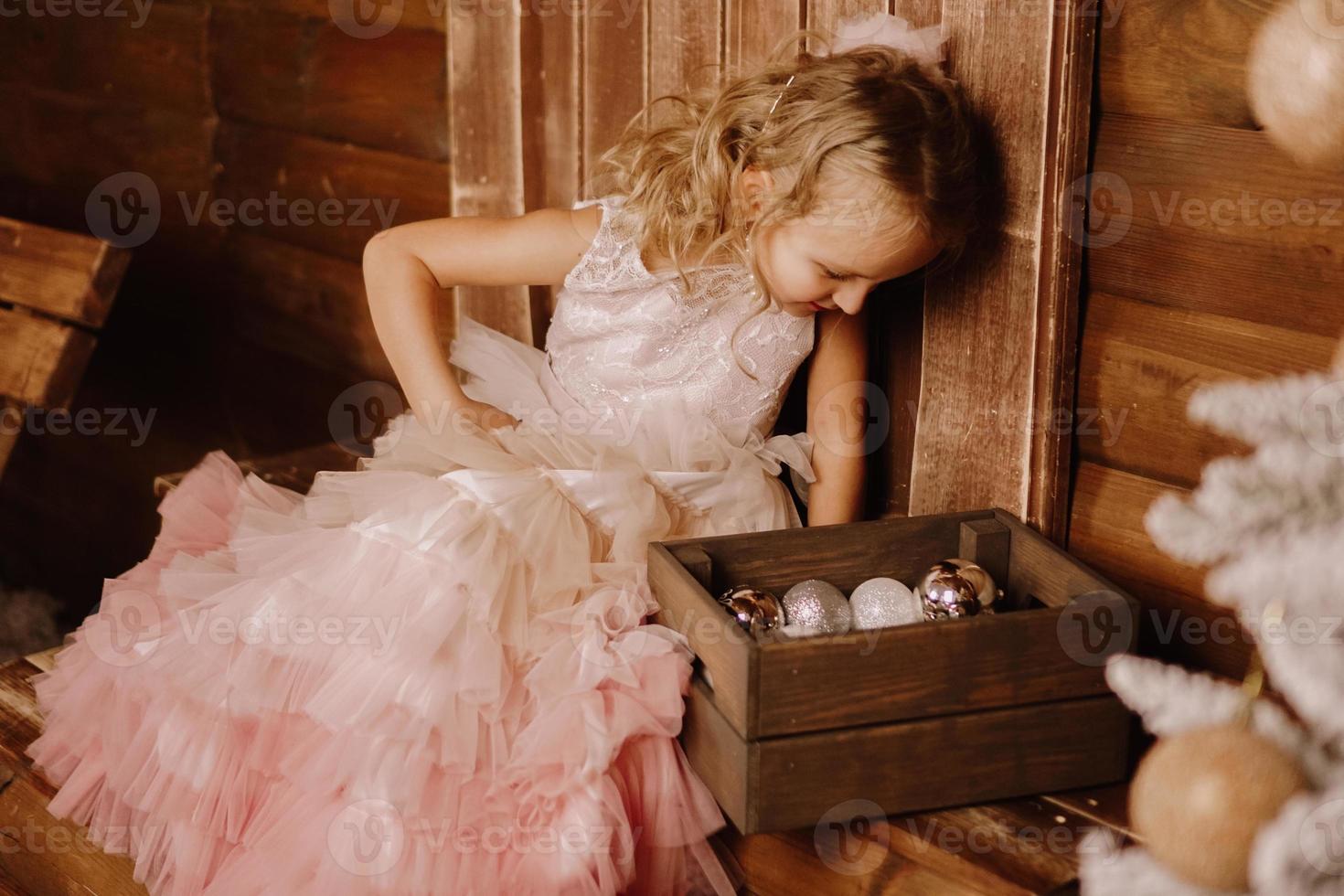 süßes Mädchen in einem rosa eleganten Kleid mit Blick auf eine Holzkiste mit Bällen foto