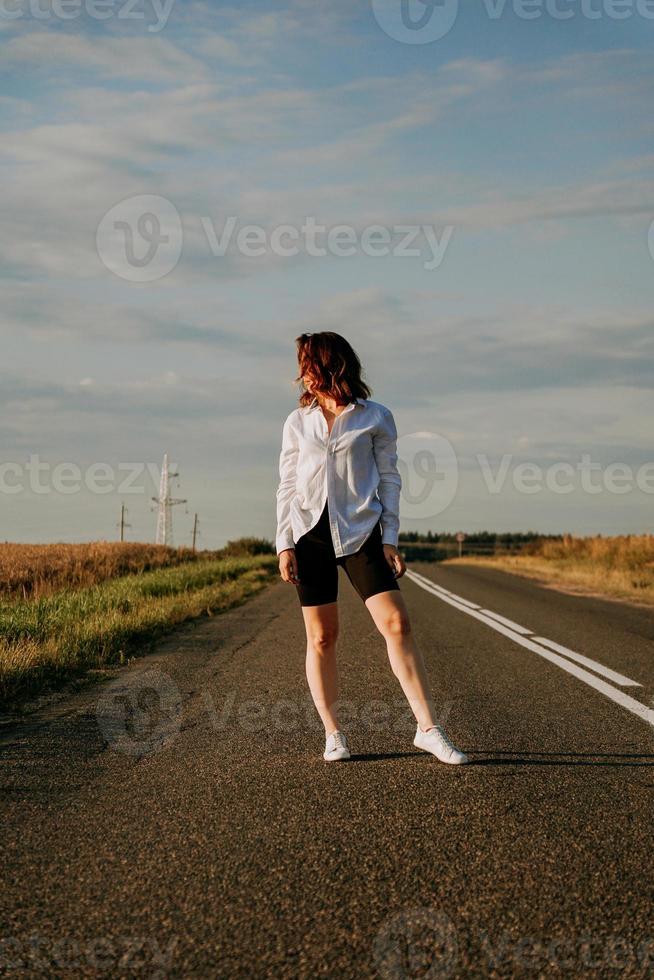 Frau in weißem Hemd geht die Straße entlang zwischen den Feldern foto