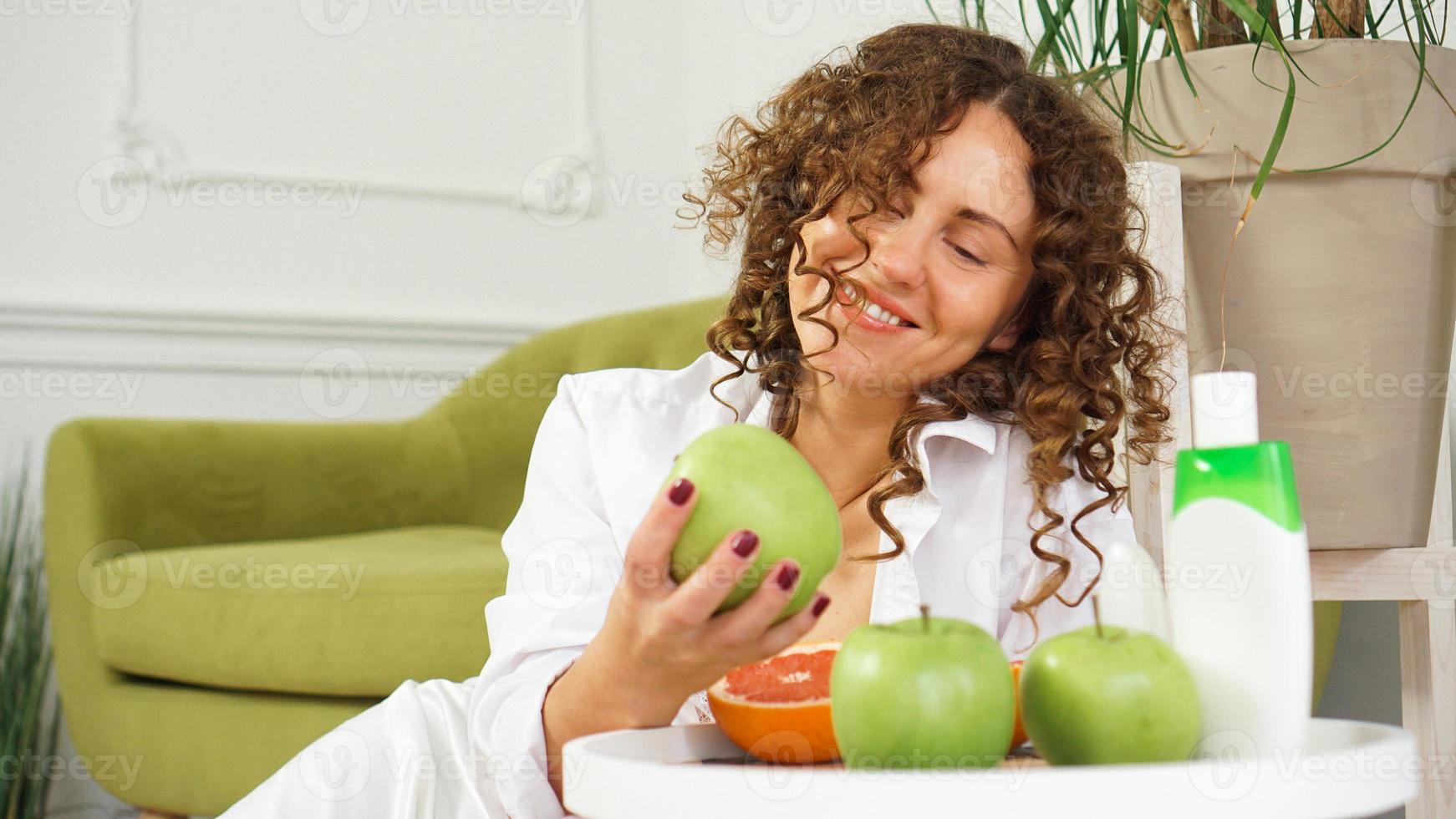 Frau mit lockigem Haar im Zimmer mit grünem Sofa. Äpfel und Bio-Pflege foto