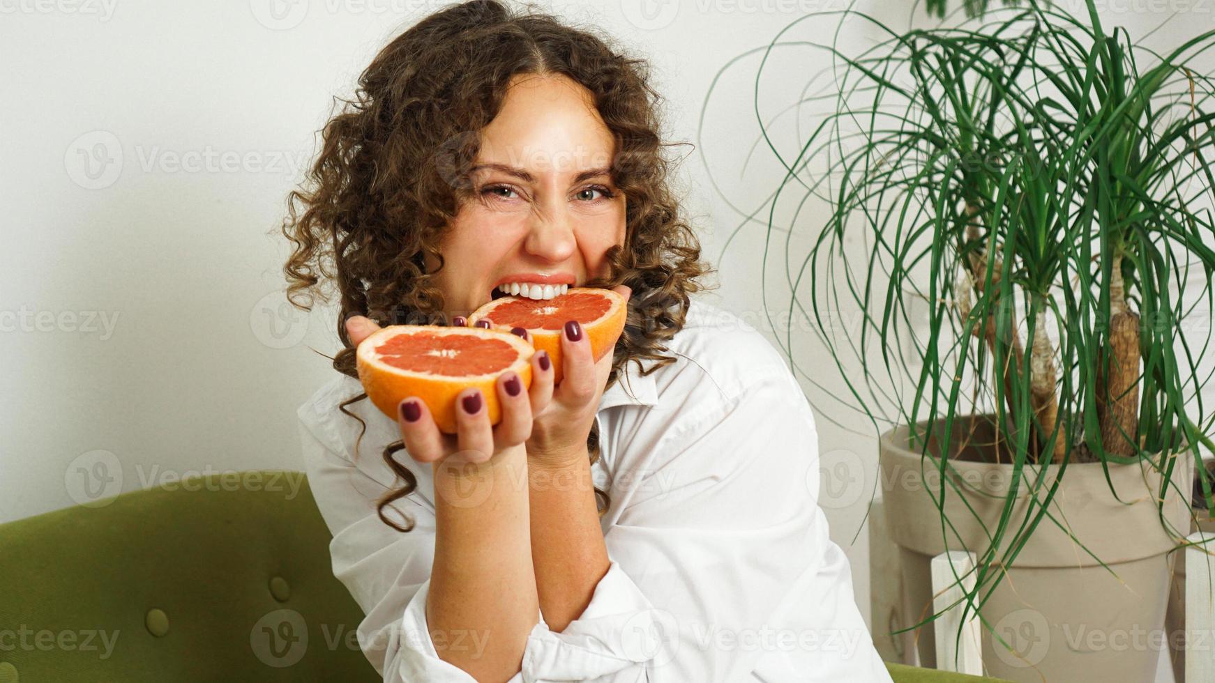 Frau beißt Grapefruit. Frau mit lockigem Haar mit Grapefruit zu Hause foto