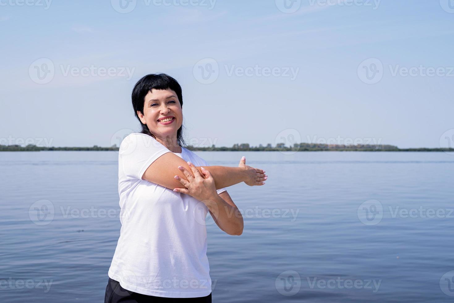 aktive und glückliche ältere Frau, die sich in der Nähe des Flussufers ausdehnt foto