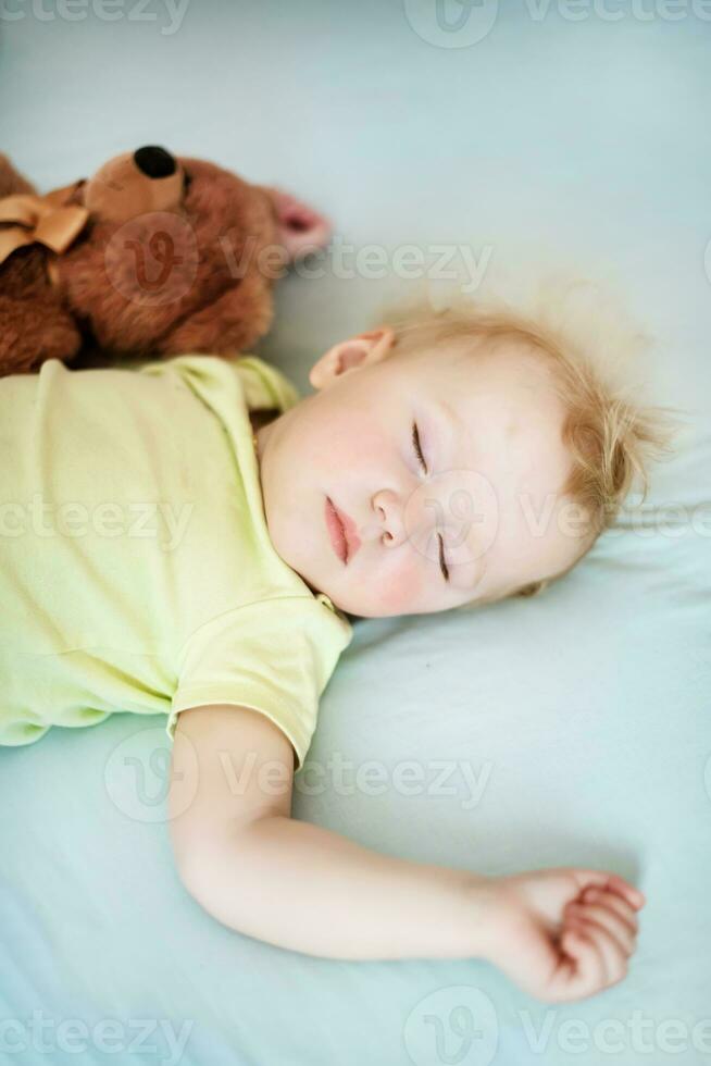 klein Kind schläft im Bett mit Teddy tragen. zerzaust Kind mit blond Haar Lügen Schließen Augen im Bett auf Blau Blatt. foto