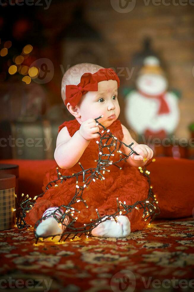 wenig Mädchen im rot Kleid gegen Hintergrund von Weihnachten Baum hält Weihnachten Girlande im ihr Hände. Baby 6 Monat alt feiert Weihnachten. foto