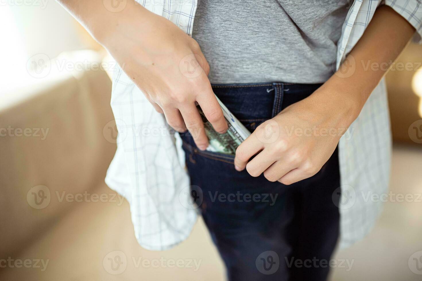 ein Teenager setzt ein Stapel von Hundert-Dollar Rechnungen im das Tasche von Jeans. ein Junge im ein Hemd versteckt sich Kasse Währung im seine Hose Tasche foto