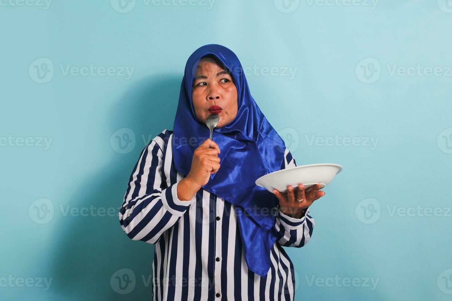 ein nachdenklich mittleren Alters asiatisch Frau im ein Blau Hijab und ein gestreift Hemd ist halten ein leeren Weiß Teller und ein Löffel während suchen zu das Seite Exemplar. sie ist isoliert auf ein Blau Hintergrund foto
