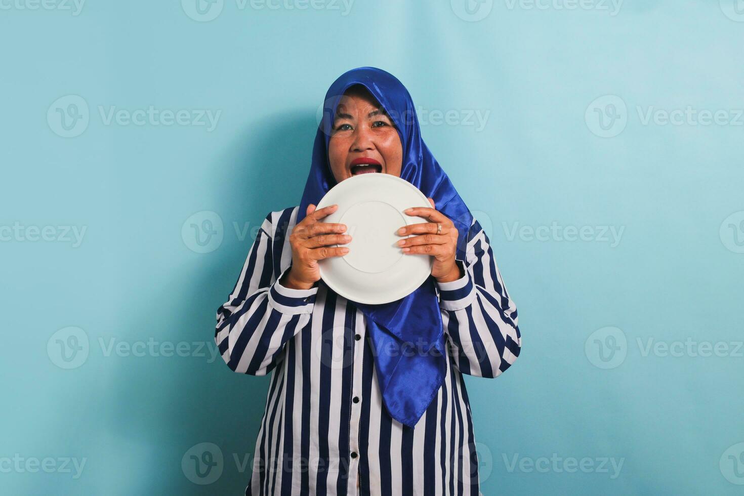 ein Süss und komisch mittleren Alters asiatisch Frau im ein Blau Hijab und ein gestreift Hemd leckt ihr Platte, immer noch hungrig nach Essen. sie ist isoliert auf ein Blau Hintergrund foto
