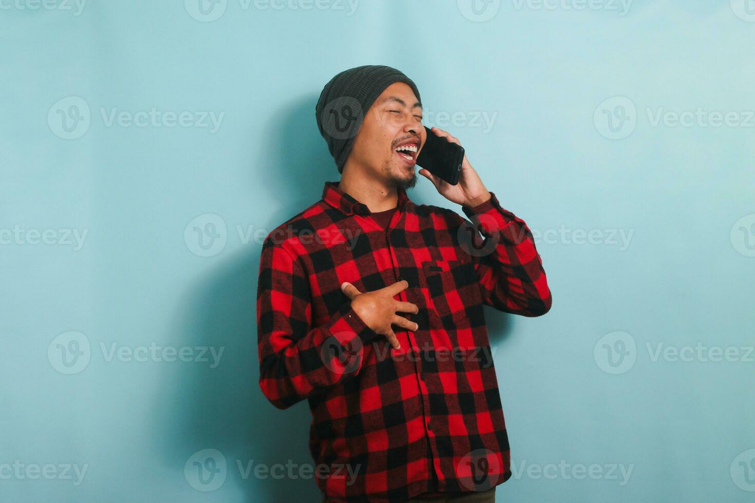 glücklich jung asiatisch Mann mit Mütze Hut und rot Plaid Flanell Hemd ist Lachen während reden auf seine Smartphone, haben ein gut Gespräch, Hören komisch Witze, isoliert auf ein Blau Hintergrund foto