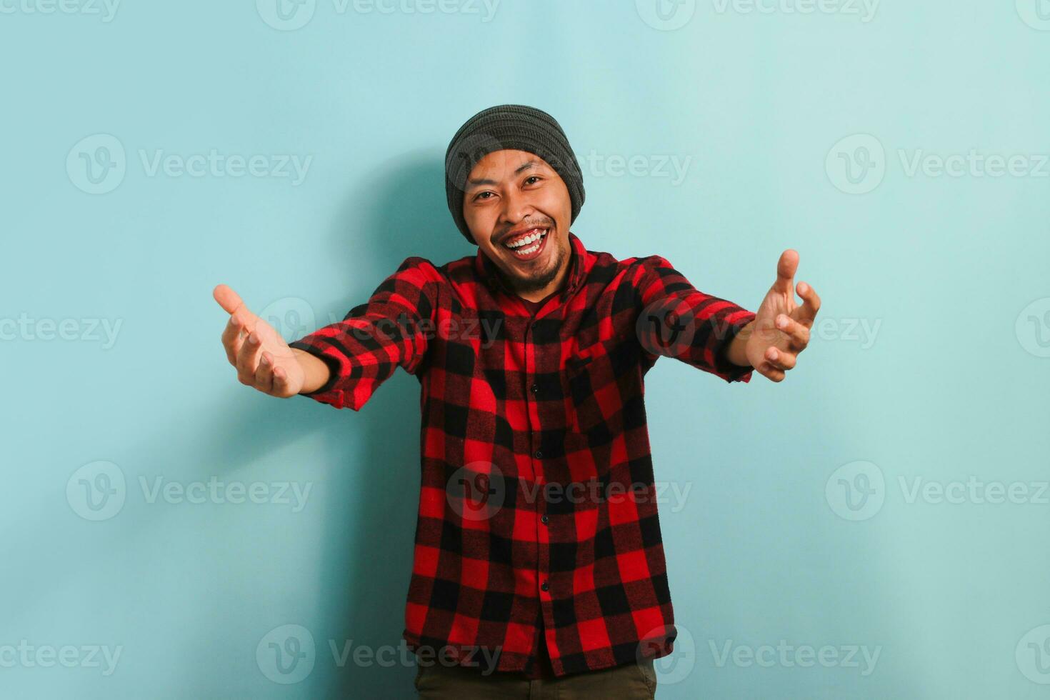 lächelnd jung asiatisch Mann mit ein Mütze Hut und ein rot Plaid Flanell Hemd erstreckt sich seine Hände gegenüber das Kamera, einladend ein Umarmung, während Stehen gegen ein Blau Hintergrund foto