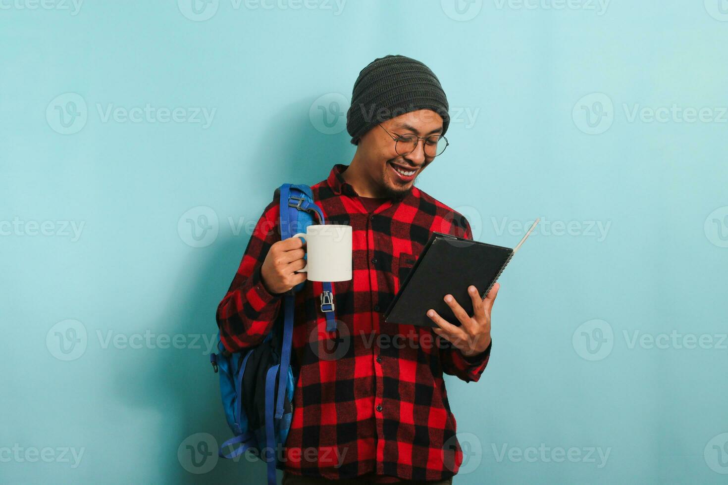 ein jung asiatisch männlich Schüler mit ein Mütze Hut und ein rot Plaid Flanell Shirt, tragen ein Rucksack und Brille, nimmt ein Kaffee brechen nach Vorträge während Stehen gegen ein Blau Hintergrund foto