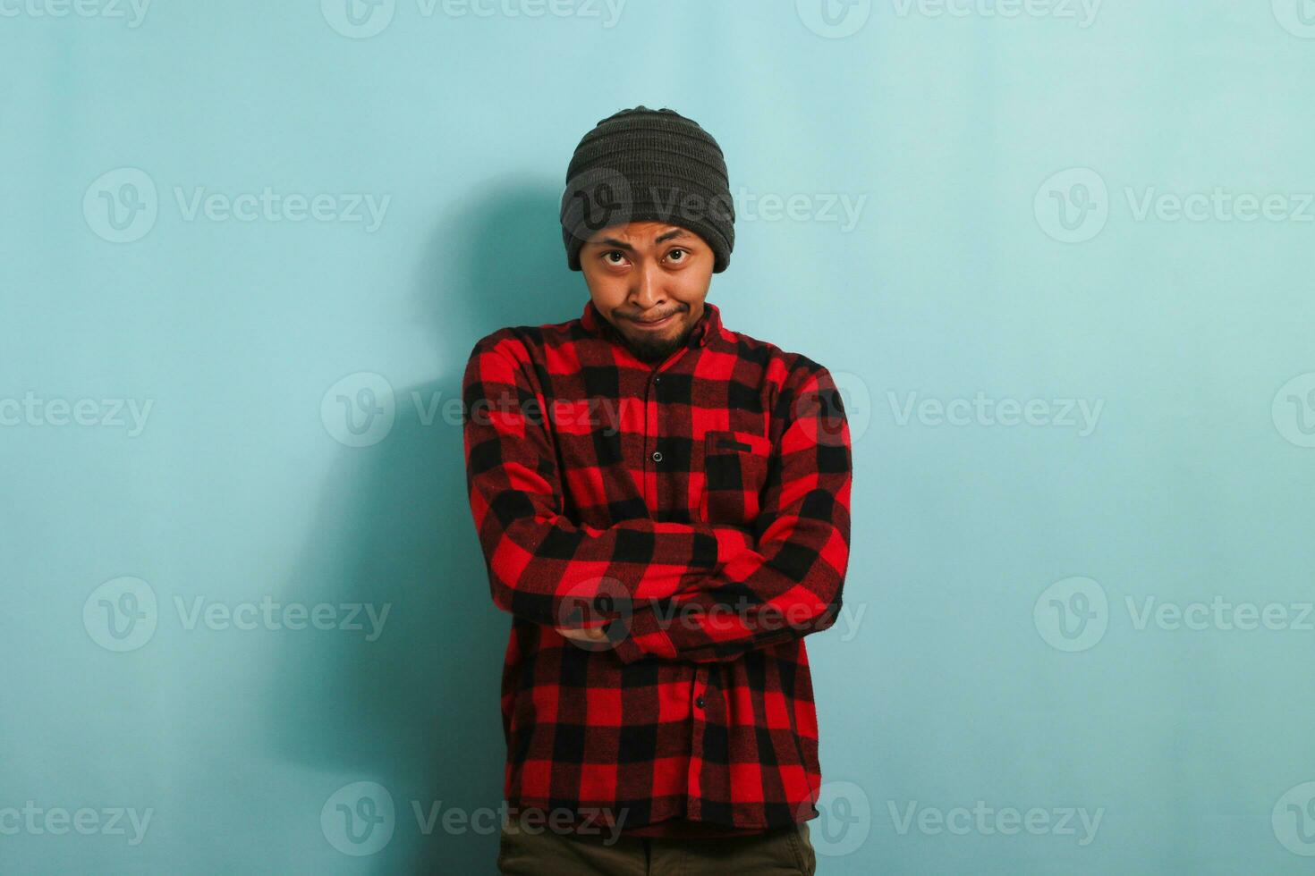 ein jung asiatisch Mann, tragen ein Mütze Hut und ein rot Plaid Flanell Shirt, erscheint verwirrt und unsicher Über ein Lösung während Stehen gegen ein Blau Hintergrund foto