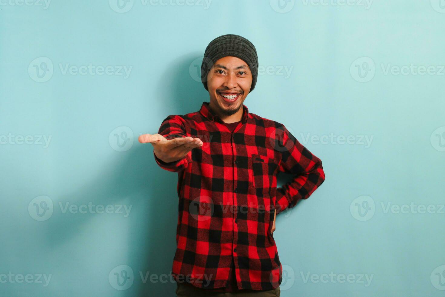 ein lächelnd jung asiatisch Mann mit ein Mütze Hut und ein rot Plaid Flanell Hemd erweitert seine Hand zum ein Handschlag, Begrüßung und Angebot Zusammenarbeit während Stehen gegen ein Blau Hintergrund foto