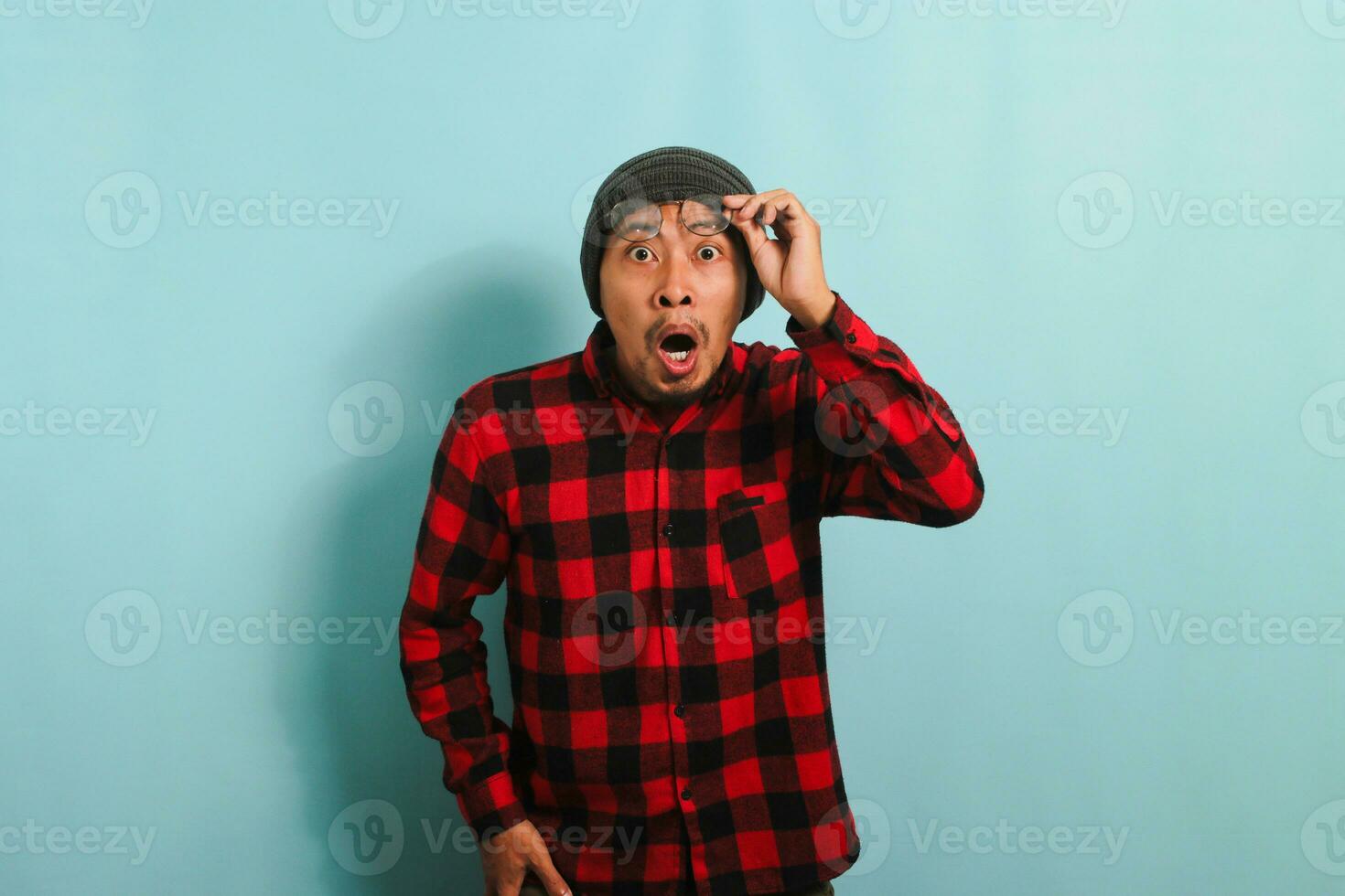 schockiert jung asiatisch Mann mit ein Mütze Hut und rot Plaid Flanell Hemd erhöht seine Brille von seine Augen im Reaktion zu unerwartet Nachricht, suchen beim das Kamera, isoliert auf ein Blau Hintergrund foto