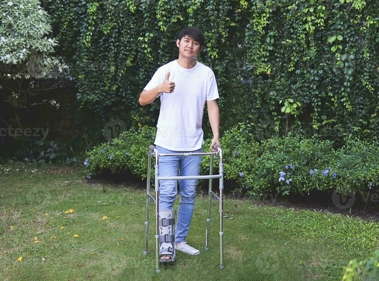 asiatisch jung Mann tragen Luftübertragung Stiefel Gehen im das Garten mit orthopädisch Gehhilfe. zeigen Daumen oben und suchen beim Kamera. foto