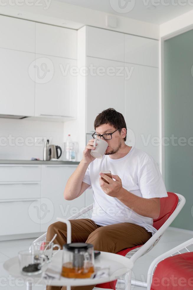 Mann sitzt auf einem Stuhl und schaut fern und hält Teetasse und Telefon in der Hand foto