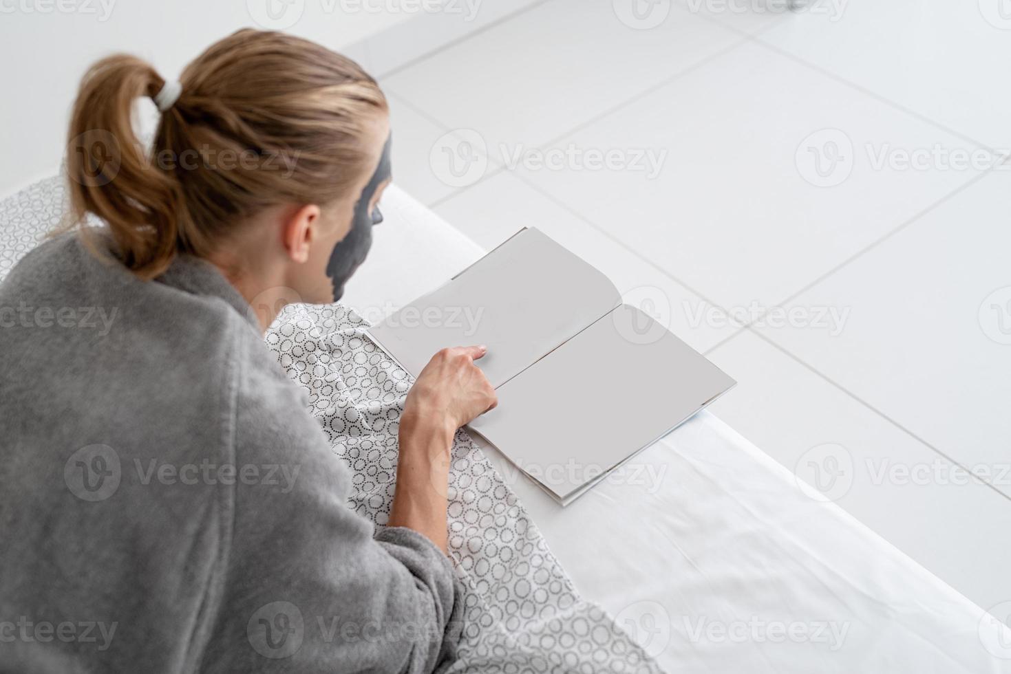 Frau mit Gesichtsmaske, die entspannt auf dem Bett liegt und eine Zeitschrift liest foto