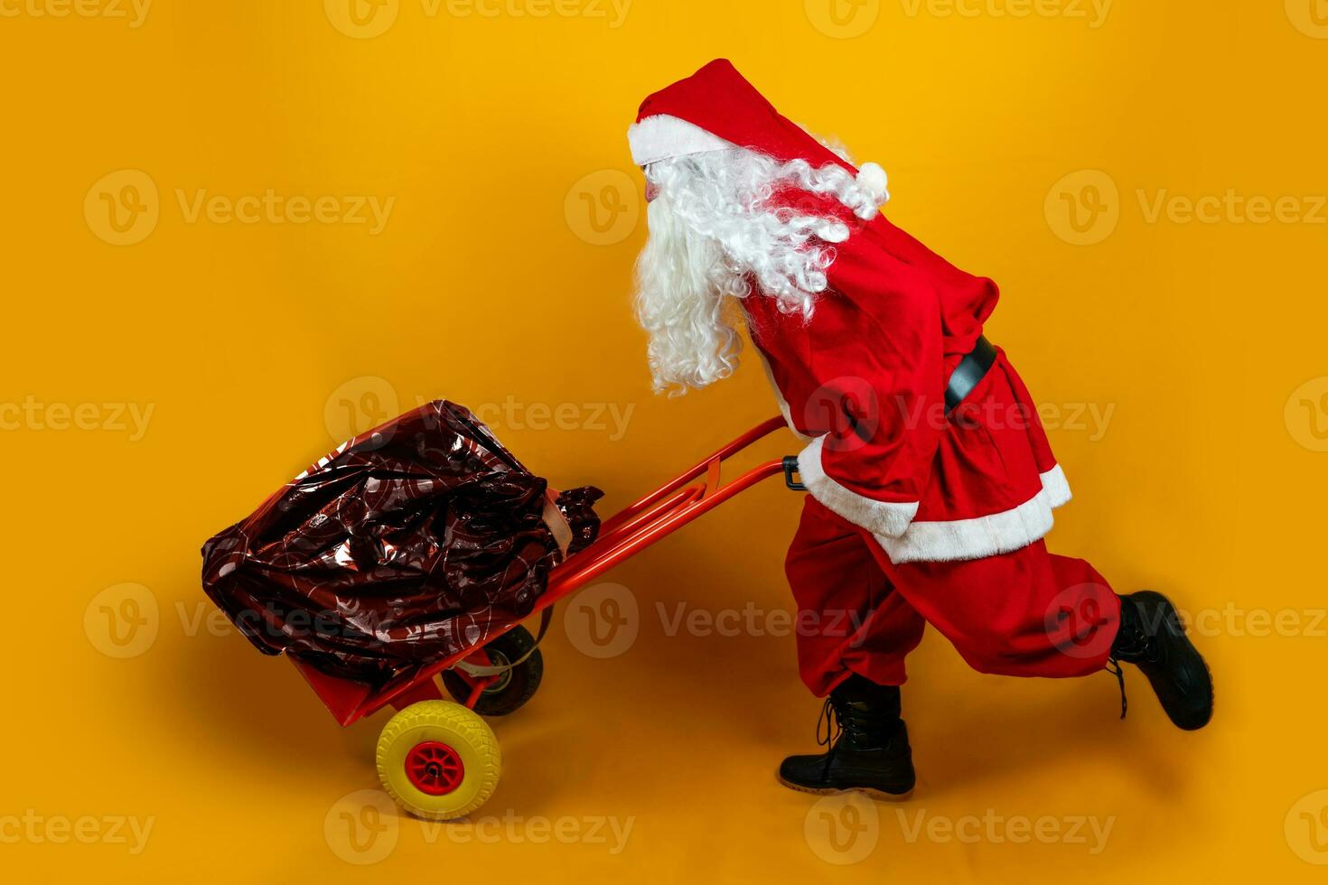 Santa claus ist Laufen mit ein Sack voll von die Geschenke auf ein Metall Wagen auf Orange Hintergrund foto