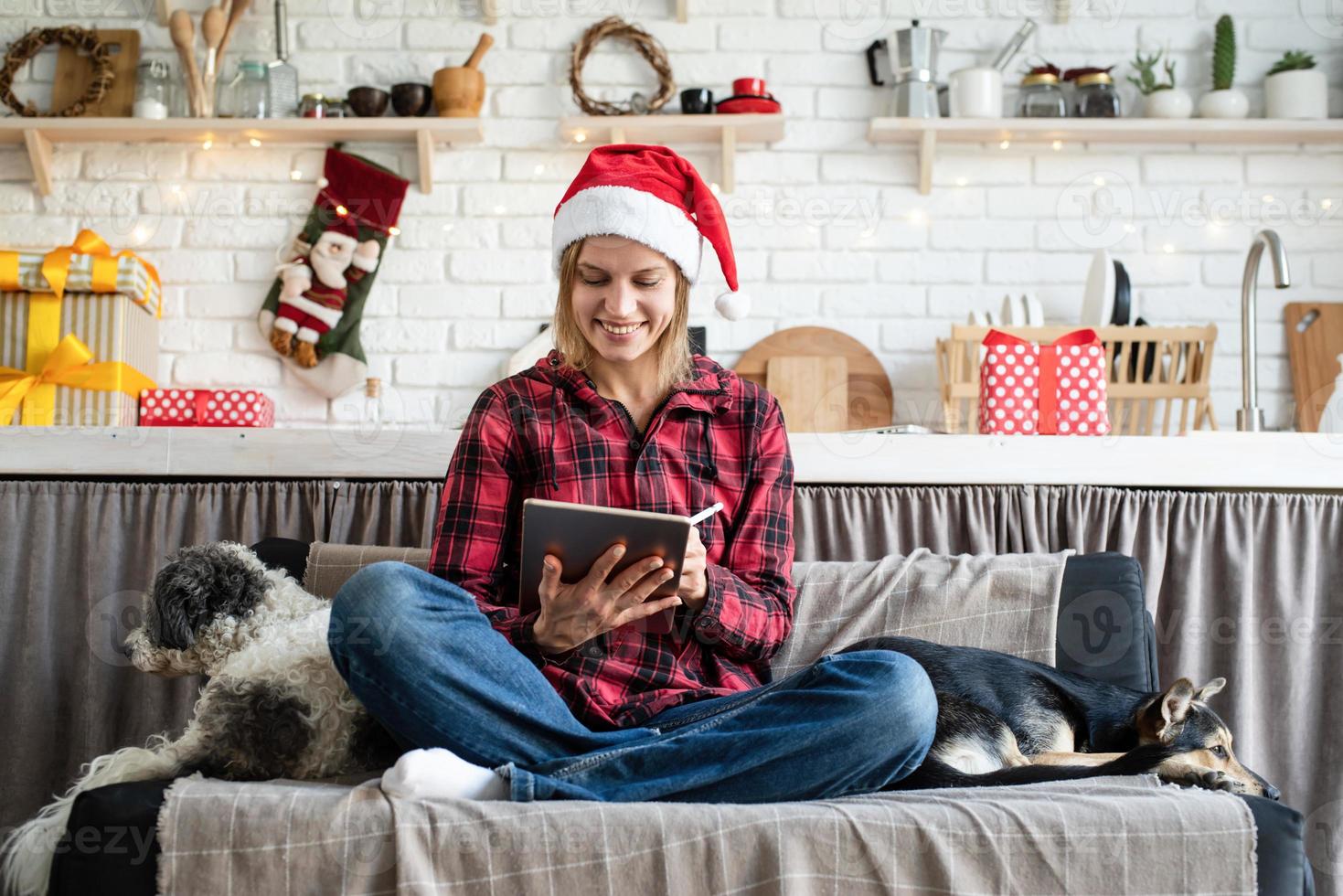 Junge blonde Frau in Weihnachtsmütze arbeitet an Tablet auf der Couch sitzend foto