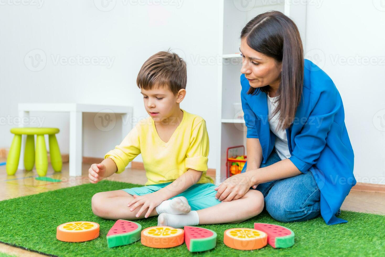 ein Frau Rede Therapeut Angebote mit ein Junge und führt aus ein Übung zu richtig das Rede Gerät durch spielen auf das Fußboden foto