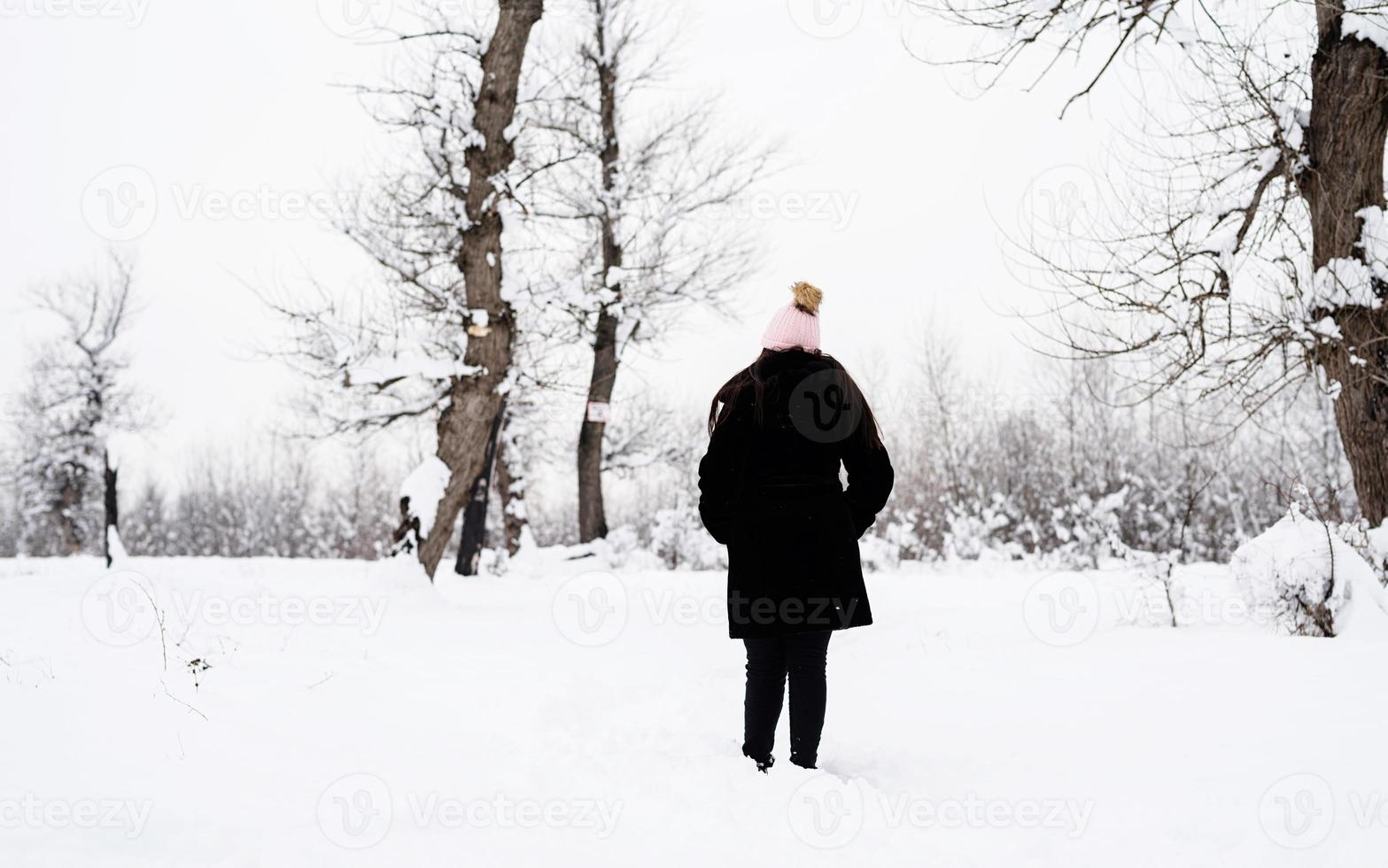 Rückansicht einer brünetten Frau, die bei Schneefall im verschneiten Park spazieren geht foto