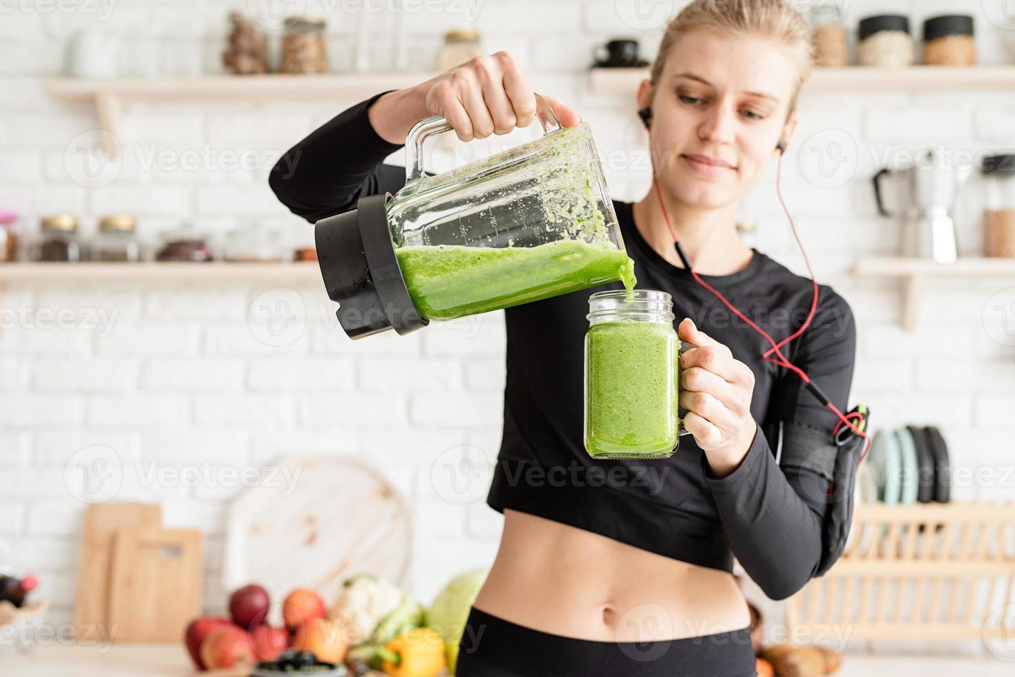 junge blonde lächelnde Frau, die zu Hause in der Küche grünen Smoothie macht foto