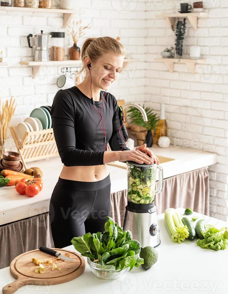 junge blonde lächelnde Frau, die zu Hause in der Küche grünen Smoothie macht foto