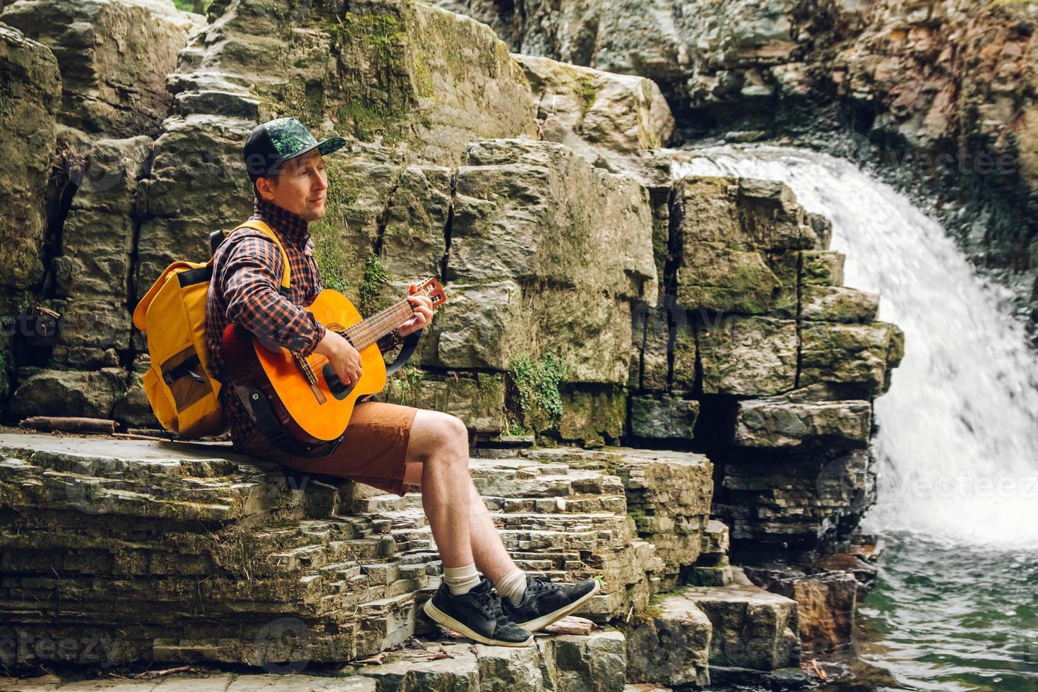 Mann spielt Gitarre, sitzt an einem Wasserfall foto