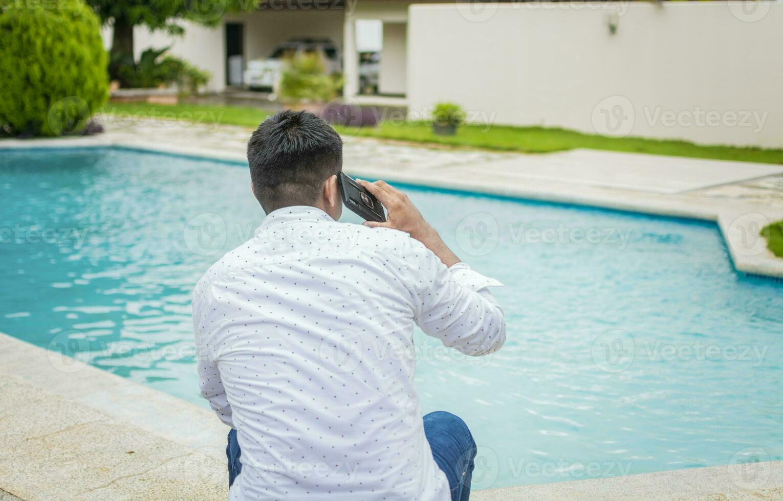 gut aussehend Mann Sitzung Berufung auf das Telefon in der Nähe von das Schwimmen Schwimmbad, jung Mann Sitzung Berufung auf das Telefon durch ein Schwimmen Schwimmbad, Kerl Sitzung reden auf das Zelle Telefon in der Nähe von das Kante von das Schwimmen Schwimmbad foto