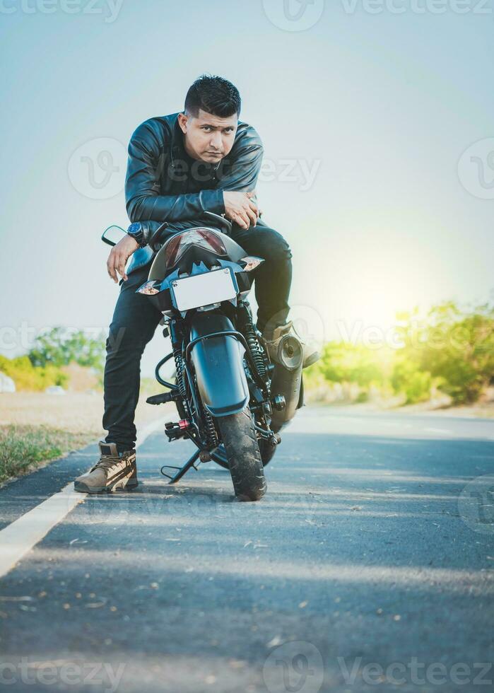 gut aussehend Motorradfahrer im Jacke Sitzung auf seine Motorrad beim das Seite von das Straße. Porträt von gut aussehend Biker auf seine Motorrad suchen beim Kamera draußen foto