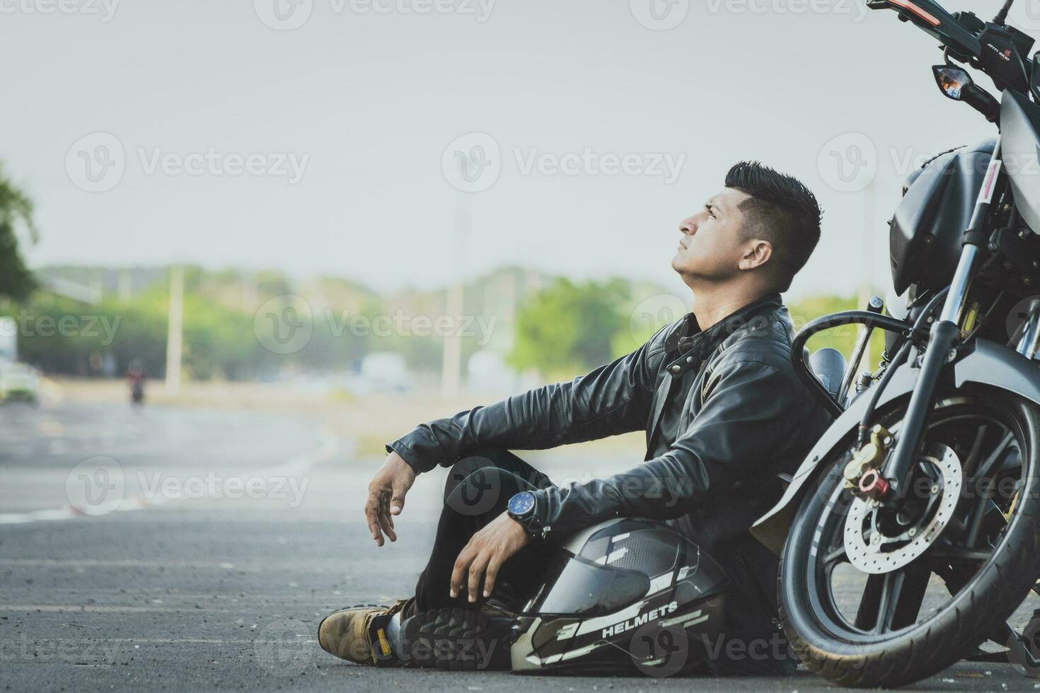 männlich Motorradfahrer Sitzung und gelehnt auf seine Motorrad auf das Asphalt. Biker Sitzung Nächster zu seine Motorrad auf das Straße foto