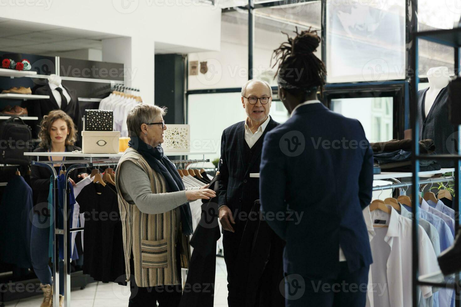 Alten Paar suchen zu Kaufen formal tragen, fragen Geschäft Manager Über Kleider Stoff im modern Boutique. Senior Mann Kauf modisch Fan-Shop und stilvoll Zubehör im Einkaufen Center foto