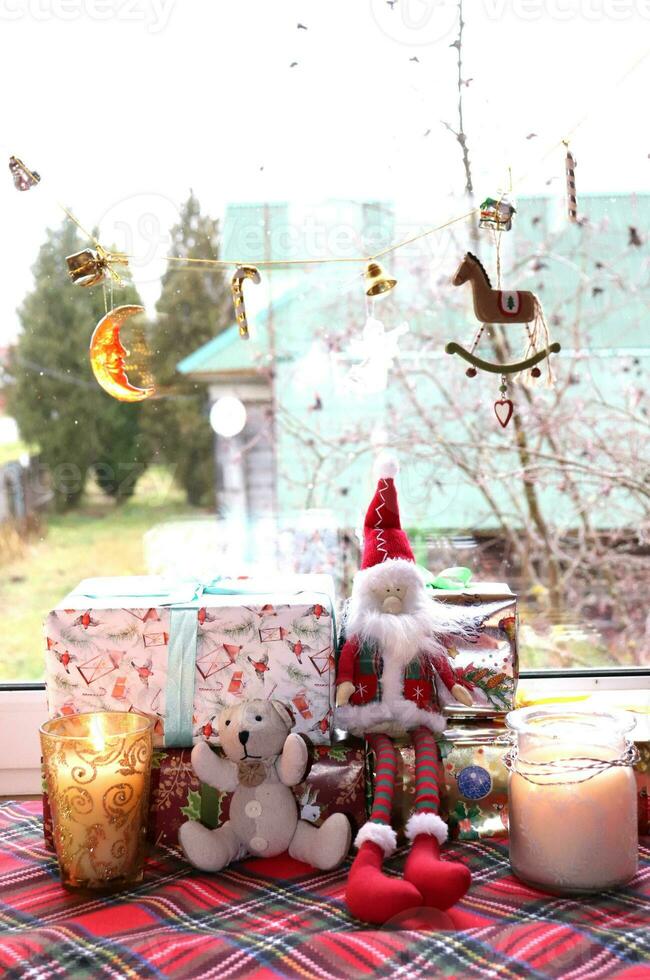 Spielzeug Gnom mit ein Bär Jungtier mit Weihnachten Geschenke und Kerzen im Glas Kerzenhalter in der Nähe von das Fenster im ein ländlich Haus, Vertikale Foto