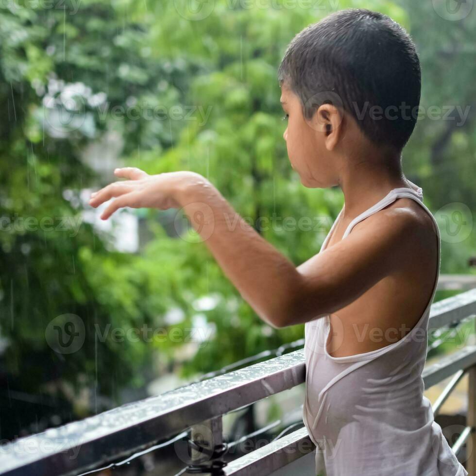 wenig Kind spielen im Sommer- Regen im Haus Balkon, indisch Clever Junge spielen mit Regen Tropfen während Monsun regnerisch Jahreszeit, Kind spielen im Regen foto