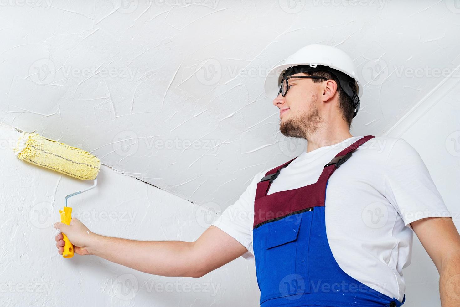 junger Mann in blauen Overalls und weißer Schutzhelm-Malwand foto