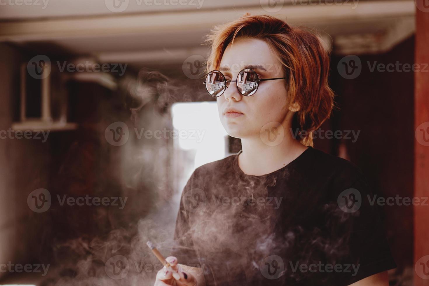Mädchen mit kurzen roten Haaren und Spiegelsonnenbrille, die Zigarette raucht foto