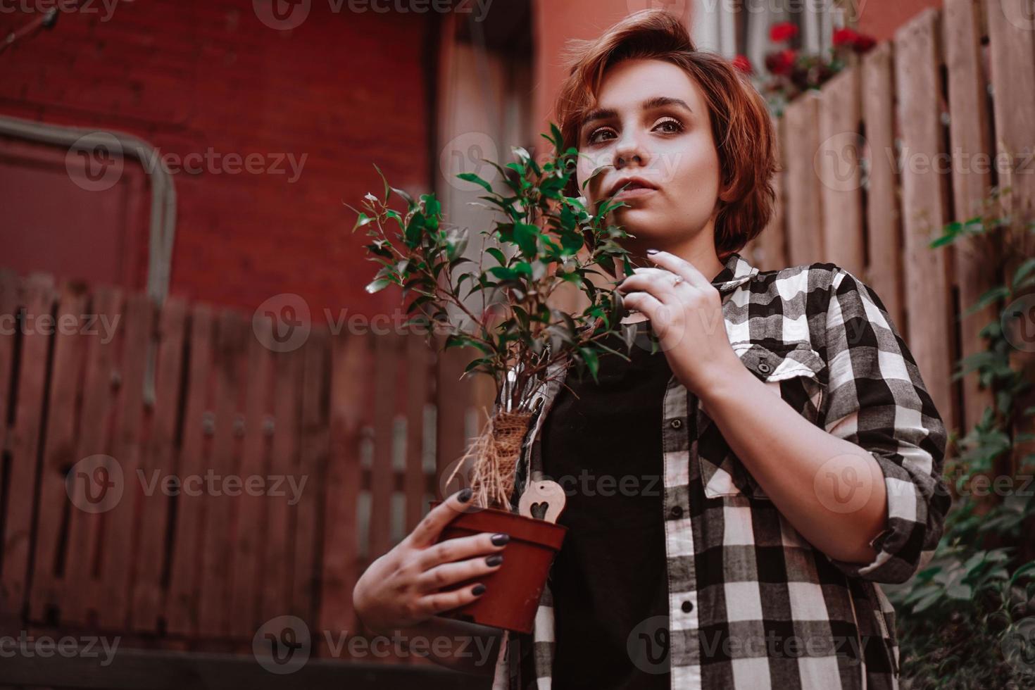 junge Frau mit kurzen roten Haaren im karierten Hemd, die eine Blume im Topf hält foto