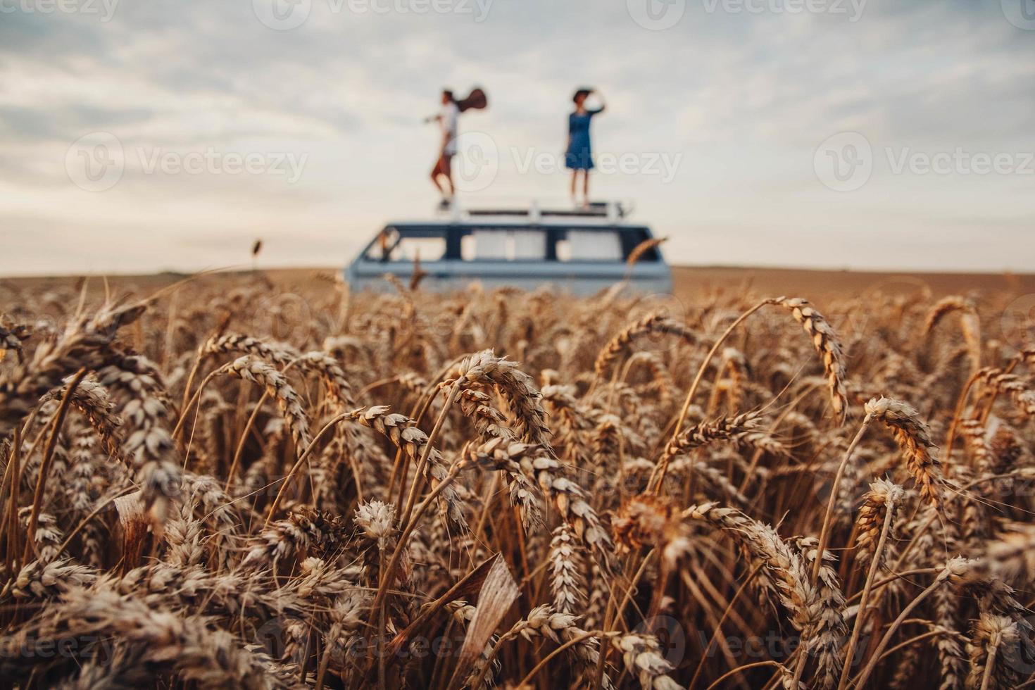 Mann mit Gitarre und Frau stehen auf dem Dach eines Autos in einem Weizenfeld foto