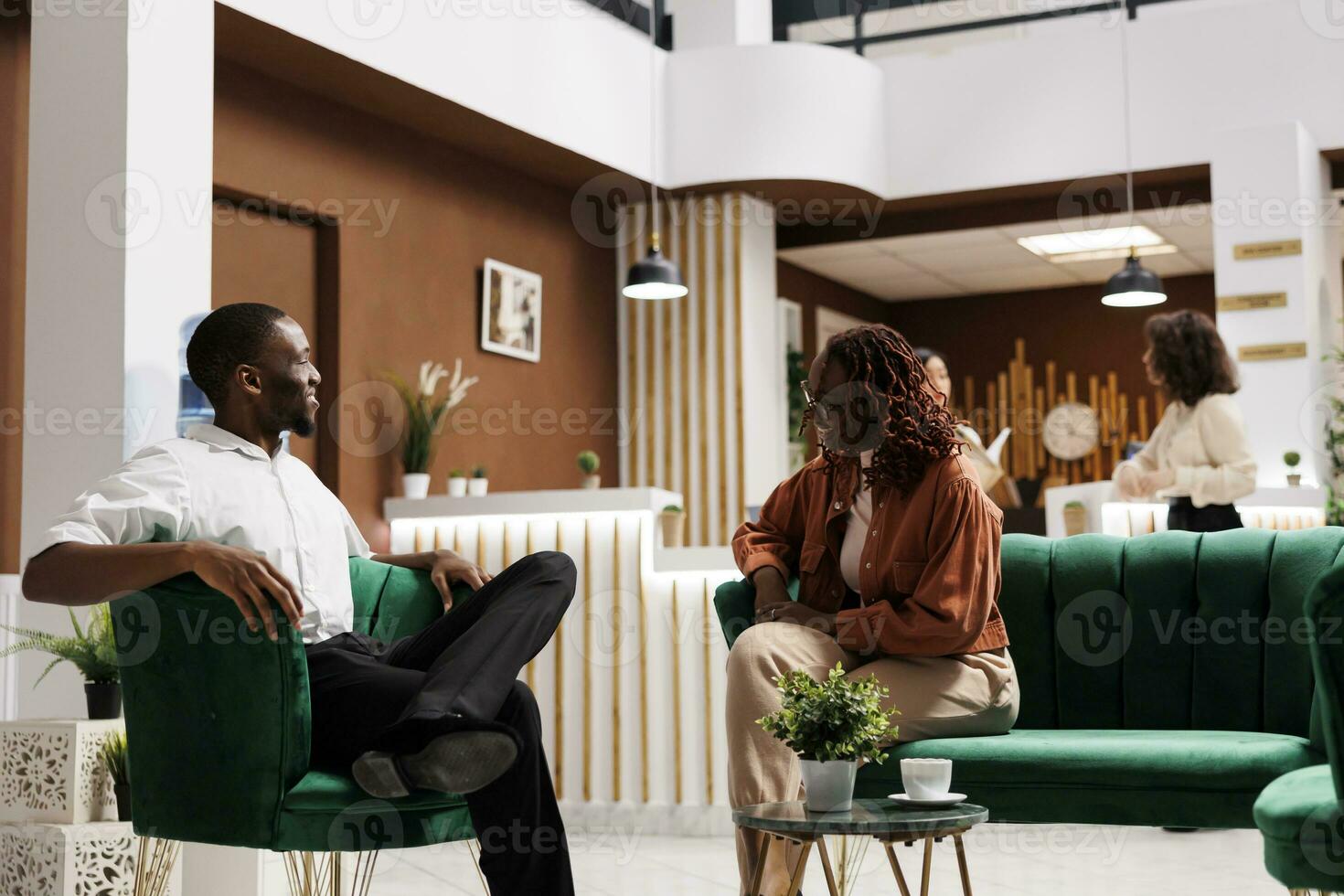 afrikanisch amerikanisch Paar reden im Empfangshalle, Sitzung im Hotel Salon Bereich und warten zu registrieren beim Rezeption Schreibtisch. glücklich Mann und Frau ankommen beim Luxus Erholungsort, entspannend auf Couch. foto