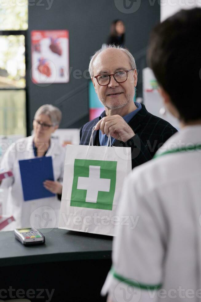 alt Mann nehmen Einkaufen Tasche von Apotheke Kassierer beim Kasse, lächelnd zu Drogerie Mitarbeiter. Alten Kunde Kauf Rezept Medikamente im medizinisch Verkauf Geschäft und Stehen beim Zähler Schreibtisch foto