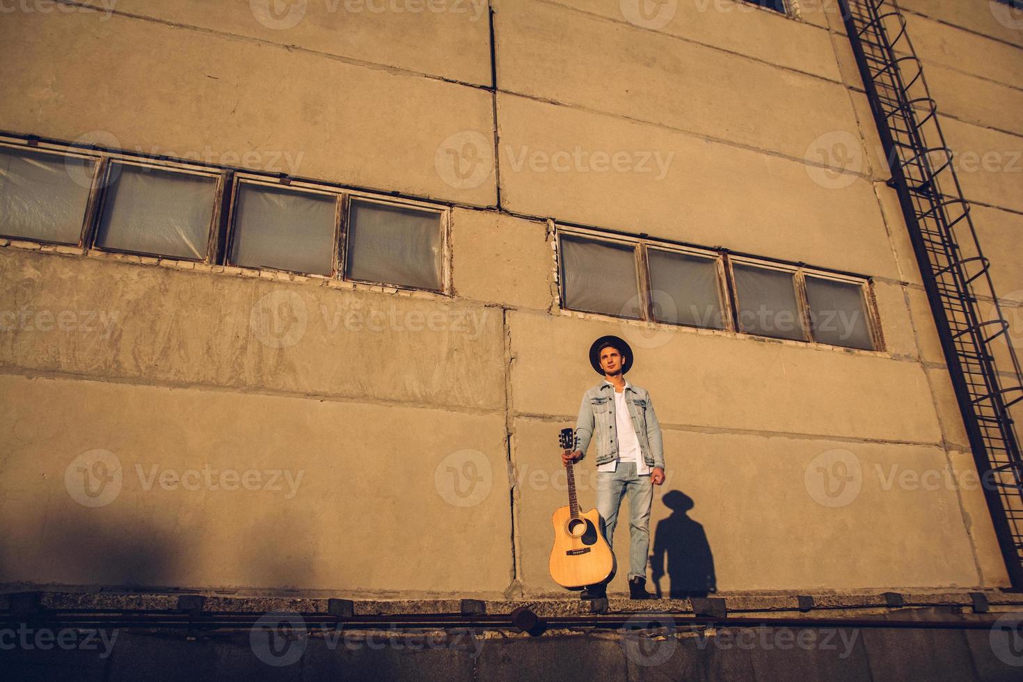 junger Kerl Musiker in einem Hut mit einer Gitarre gegen eine Betonwand foto