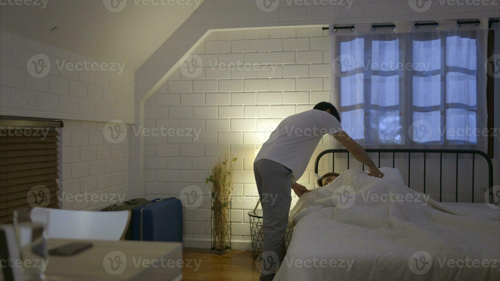 ein jung asiatisch Mann Arbeiten im Laufe der Zeit beim Zuhause Abdeckungen das Decke zum seine Schlafen Ehefrau auf das Bett. vor zu auch weiterhin Arbeit foto