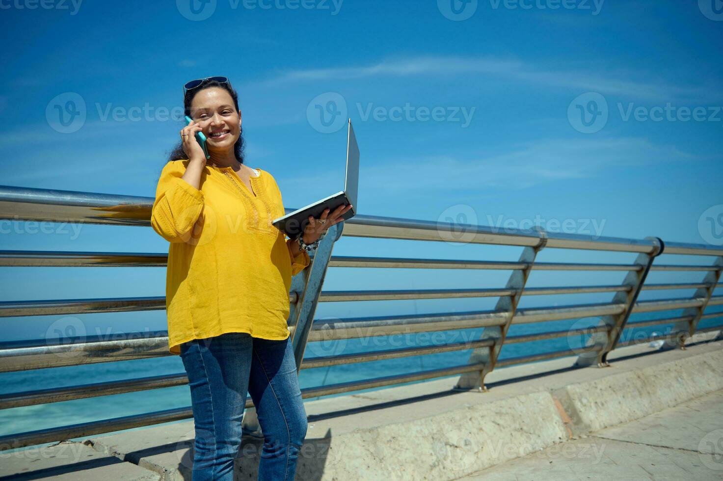 zuversichtlich Multitasking Der Umsatz Manager hält ein Laptop, online funktioniert und lächelt während ein Konversation auf Clever Handy, Mobiltelefon Telefon foto